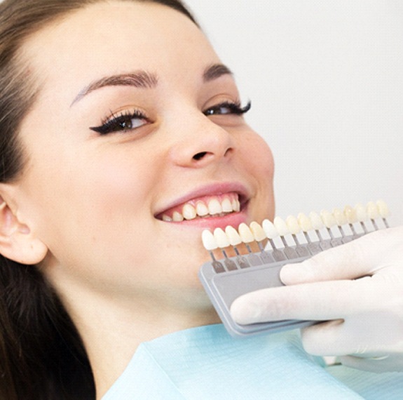 A young female sits back in a dentist’s chair while a dental professional uses a shade guide to determine the color of her veneers