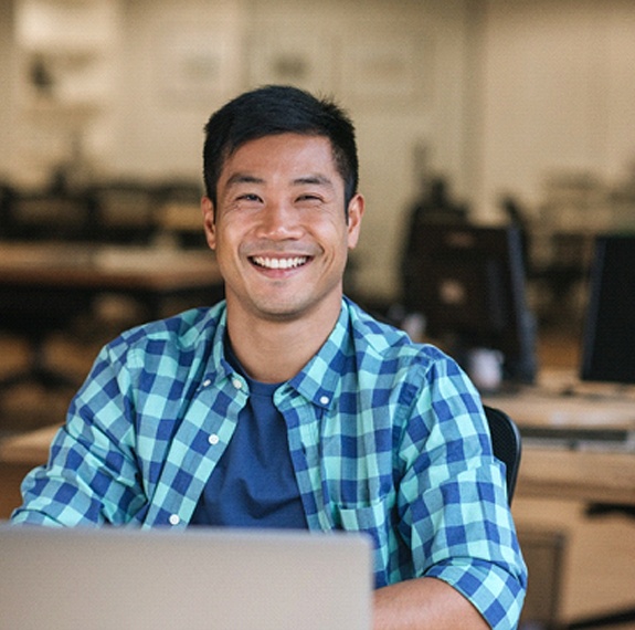 A young man wearing a blue and green plaid button-down shirt smiles while seated at a computer