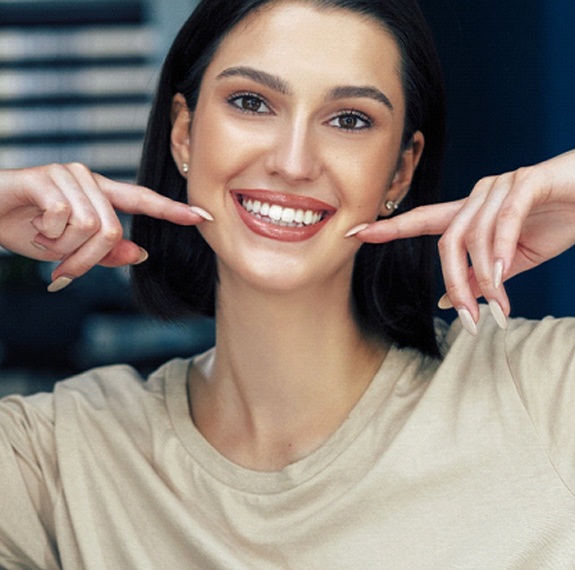 A young woman with dark hair smiles wide to show off her veneers while pointing to her mouth