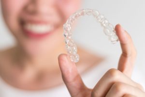 Blurry image of woman over white background holding up Invisalign with 2 fingers in the foreground
